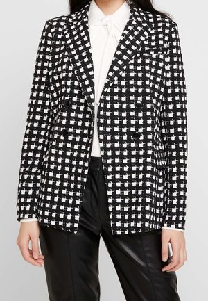 Zwart-wit geruite dubbelgeknoopte blazer met vier knopen, gedragen over een witte blouse en zwarte leren broek.