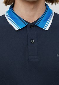 Marinblå polotröja med texturerad tyg, utrustad med en ljusblå och vit randig krage samt en svart knapp vid halslinningen.