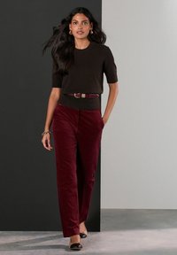 Brązowy dopasowany sweter z krótkim rękawem, bordowe szerokie spodnie z kordurowej tkaniny i czarne baleriny. Modelka stoi pewnie na neutralnym tle.