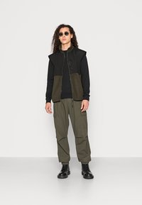 Svart zip-up vest med olivengrønn paneling over en svart skjorte, kombinert med olivengrønne cargo-bukser og svarte snøresko. Runde solbriller er inkludert.