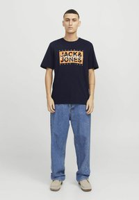 Námorně modré bavlněné tričko s oranžovým grafickým nápisem "JACK & JONES". V kombinaci s volnými modrými džíny a béžovými botami. Jednoduchý design a volný střih.