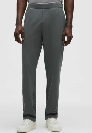 Moški v antracitno sivih hlačah s ravnimi nogavicami in belih športnih copatih, stoji ob preprostem svetlem ozadju.