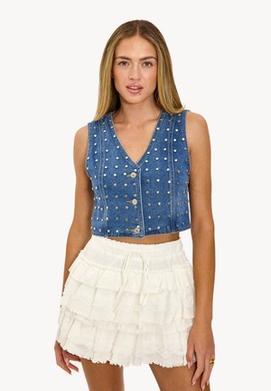 Gilet en denim avec un design en V, featuring des clous argentés, et une longueur courte, associé à une jupe blanche à volants superposés.
