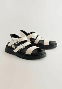 Sandali con cinturini, suola in gomma nera, cinturini in sintetico bianco e fibbie in metallo. Design con punta aperta e tacco piatto.