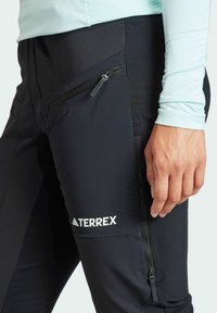 Svarta Terrex-byxor med slim fit, som har en dragkedjeficka på låret, slät textur och en vit logotyp på benet.