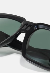 Ochelari de soare negri cu lentile verzi închise, având o ramă groasă și accente decupate. Designul subliniază o formă îndrăzneață, geometrică.
