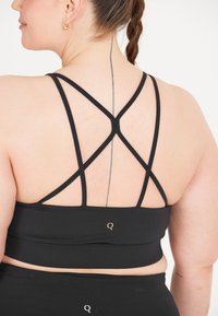 Zwarte sport beha met gekruiste bandjes, voorzien van een gladde textuur, een elastische band en een klein goud logo op de achterkant.