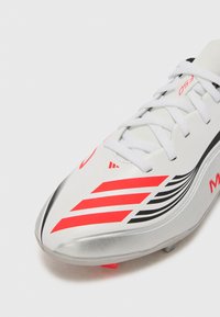 Crampons de soccer blancs avec une semelle argentée brillante, comportant des motifs de rayures noires et rouges et des lacets blancs. Design robuste pour un usage athlétique.