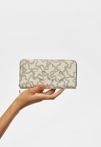 Beige wallet with a textured pattern of wavy lines in darker tones, featuring a zip closure and logo detail embossed on the front.