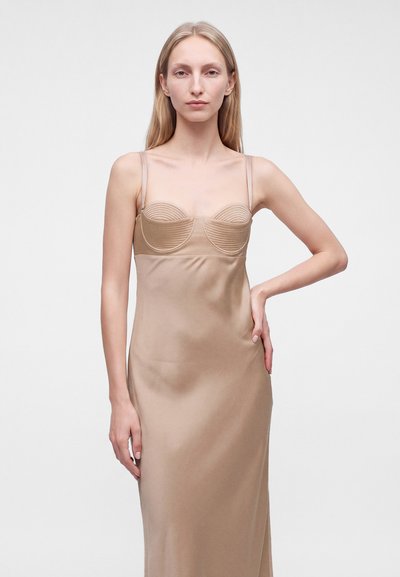 Femme aux longs cheveux blonds, portant une robe nuisette en satin beige à fines bretelles, debout devant un fond clair uni, une main sur la hanche.