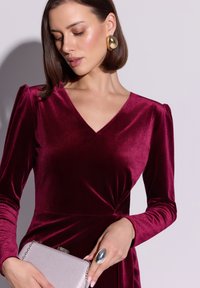 Burgundowa sukienka z aksamitu z dekoltem w serek, bufiastymi rękawami i skręcanym detalem w talii. Srebrna kopertówka i duży pierścionek z akcentami złota.