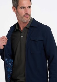 Marine blauwe ritsjack van een gestructureerde stof met een borstzak, gedragen over een donkergroene polo met een fijn meshpatroon. De binnenkant heeft een blauwe print.
