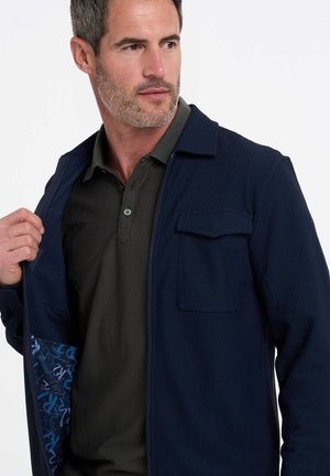 Marine blauwe ritsjack van een gestructureerde stof met een borstzak, gedragen over een donkergroene polo met een fijn meshpatroon. De binnenkant heeft een blauwe print.