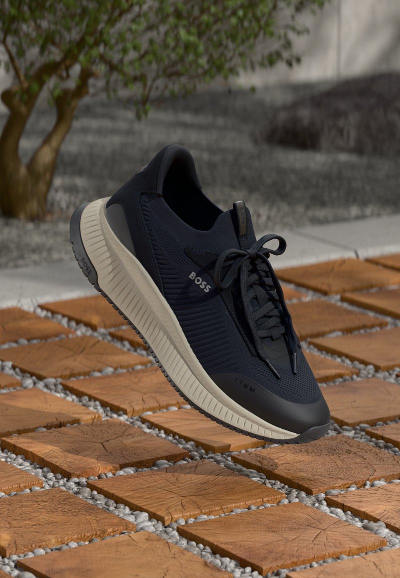 Navy breathable sneakers featuring a textured upper, lightweight design, and contrasting beige sole, set on interlocking wooden tiles.