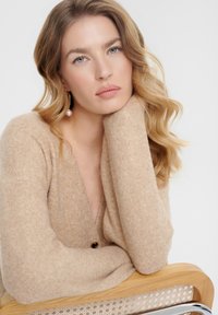Cardigan beige lavorato a maglia con scollo a V profondo, maniche lunghe e un singolo bottone nero. Texture morbida, abbinato a una sedia di legno.