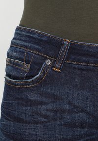 Närbild av en ficka på mörkblå denimjeans med orange sömnad, som bärs tillsammans med en olivgrön topp.