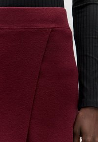 Bordeauxfarbene Strickrock mit einem eng anliegenden Taillenbund und einem schrägen Saum. Kombiniert mit einem schwarzen, strukturierten Oberteil mit langen Ärmeln. Weicher Stoff.