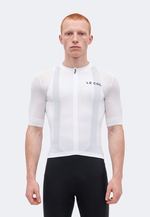 Mann mit kurzen roten Haaren, der ein eng anliegendes weißes Radtrikot mit dem "LE COL"-Logo und schwarze Radhosen trägt, steht vor einem einfarbigen Hintergrund.