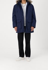 Marinblå parkas med pälskant på huvan, två framfickor och dragkedja framtill. Bärs över en vit skjorta och svarta byxor, kombinerat med vita sneakers.