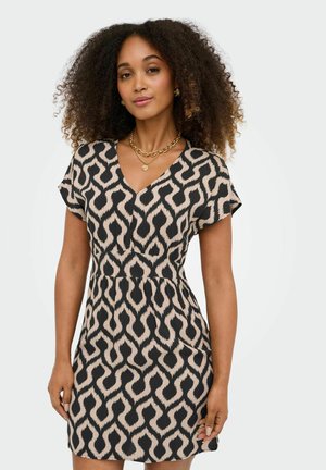 Femme aux cheveux bouclés portant une robe beige à manches courtes avec un motif géométrique noir audacieux et un collier chaîne doré sur fond uni.