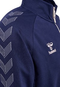 Mørkeblå zip-up sweatshirt med høj krave, der har hvide diagonale stribede chevrons på venstre arm og et logo på brystet.