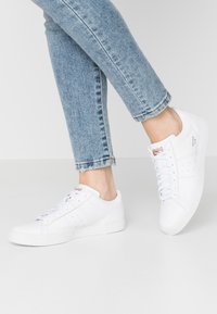 Witte leren sneakers met een gestructureerde zool, met perforaties aan de zijkanten en vetersluiting. Gekleed met lichtblauwe rafelige jeans.