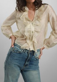 Blusa avorio traslucida con davanti arricciato, leggera lucentezza e paillettes decorative, abbinata a jeans in denim blu chiaro con silhouette aderente.