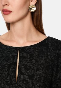 Runde, metallische goldene Ohrringe mit einer glatten, reflektierenden Oberfläche. Das Kleid besteht aus strukturierter schwarzer Stoff mit floralen Mustern und einem Schlüssellochausschnitt.