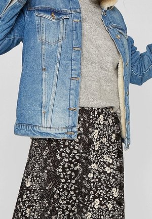 Chaqueta de mezclilla con un acabado azul claro, presenta botones plateados y dos bolsillos en el pecho, sobre un suéter gris y una falda negra con flores y botones.