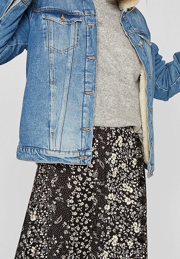 Jeansowa kurtka w jasnoniebieskim odcieniu, ozdobiona srebrnymi guzikami i dwoma kieszeniami na piersi, noszona na szarym swetrze i czarnej spódnicy w kwiaty z guzikami.