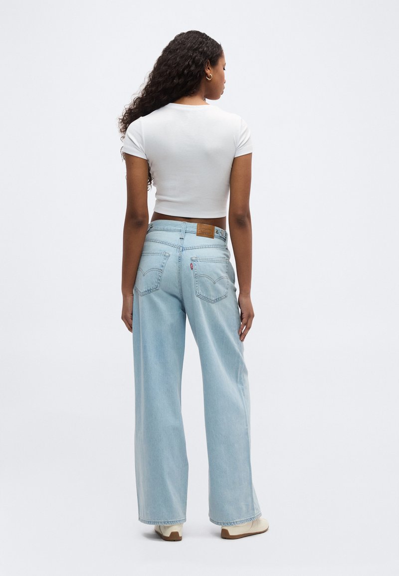 Femme aux longs cheveux bouclés de dos, portant un t-shirt blanc court, un jean ample bleu clair et des baskets blanches.