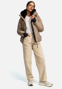 Tan gewatteerde pufferjas met zwarte nepbont kraag, ritszakken en zwarte accenten. Gecombineerd met beige wijd uitlopende broek en sneakers.