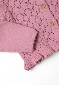 Roze gebreide cardigan met rozenmotieven van kant in diamanten vormen, houten knopen en geribde manchetten en zoom op een witte achtergrond.