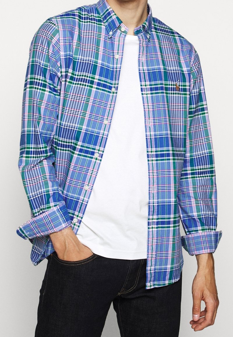 Un homme porte une chemise à carreaux bleue et verte boutonnée sur un t-shirt blanc avec un jean noir, main dans la poche, sur un fond uni.
