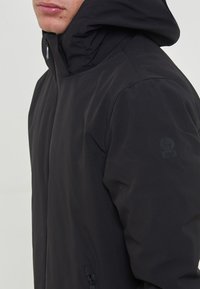 Giacca nera con cappuccio realizzata in materiale leggero, dotata di chiusura a zip, cappuccio regolabile e dettaglio del logo sulla manica.