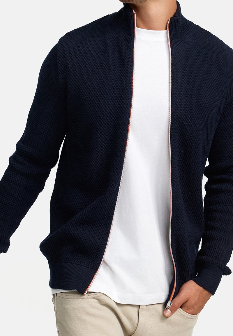Marinefarbene, strukturiert e Zip-Jacke mit hohem Kragen, kontrastierendem roten Reißverschluss und gerippten Bündchen, getragen über einem weißen T-Shirt.