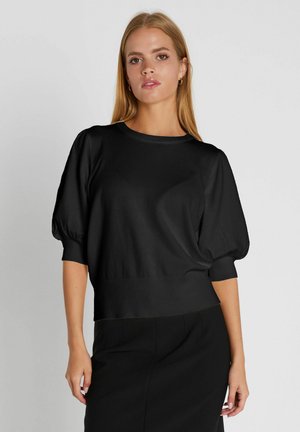 Mladá žena s dlhými blond vlasmi, ktorá má oblečený čierny top s nadýchanými rukávmi a čierne nohavice, stojaca pred jednoduchým svetlým pozadím.