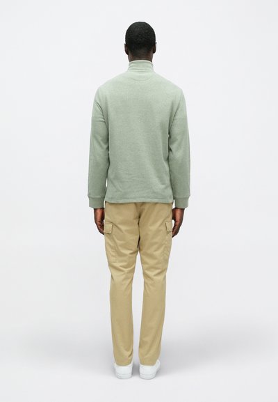 Πράσινη μπλούζα φούτερ με ψηλό γιακά και μακριά μανίκια, συνδυασμένη με ανοιχτό μπεζ cargo παντελόνι που διαθέτει πλαϊνές τσέπες. Λευκά αθλητικά παπούτσια.