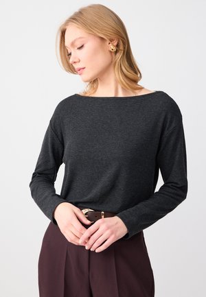 Vrouw met blond haar draagt een donkergrijze top met lange mouwen, een bruine broek, een gouden gesp en gouden oorringen, en kijkt iets naar beneden.