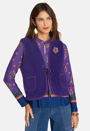 Purple fleece vest with two front pockets, tied at the waist. Worn over a multi-patterned long-sleeve shirt with floral designs.