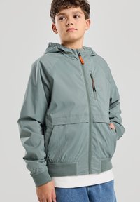 Vihreä huppujakku, jossa on vetoketju etuosa, elastiset hihansuut ja sivutaskut. Sisältää vetoketjutaskun, jossa on oranssi yksityiskohta ja sileä pinta.