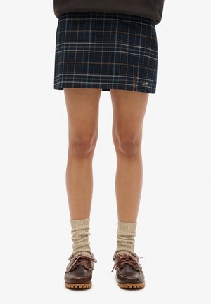 Jupe mini à carreaux marine avec des lignes orange et blanches, dotée d'une fente latérale. Associée à des chaussettes beiges et des chaussures marron à lacets.