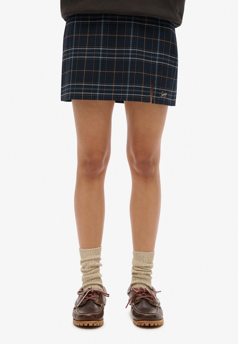 Granatowa mini spódnica w kratę z brązowymi i białymi liniami, z bocznym rozcięciem i haftowanym logo. Noszona z beżowymi prążkowanymi skarpetkami i brązowymi butami.