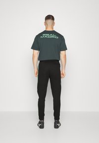 Mand i sort "Real Madrid" t-shirt, sorte joggers med grønne striber, sorte sportsko, står og vender mod hvid væg.