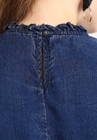 Vue arrière d'une personne portant un haut en denim bleu avec un col volé et une fermeture à boutons au cou, portant une boucle d'oreille en or.
