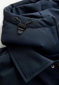 Navy blue jacket featuring a high collar, adjustable drawstring, and zipper closure. Smooth texture with visible stitching and hardware accents.