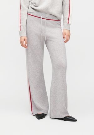 Pantalon large en maille gris clair avec bandes latérales rouges et taille à cordon, porté avec des chaussures pointues noires et un pull assorti gris clair.