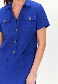 Vestito in lino blu con maniche corte, chiusura frontale con bottoni, due tasche sul petto e due tasche inferiori. Presenta quattro bottoni tondi e un colletto.