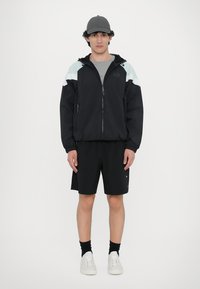Jachetă windbreaker neagră cu accente albastre deschise și albe, cu fermoar frontal, mâneci elastice, asortată cu pantaloni scurți negri și adidași albi.