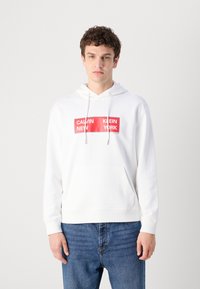 Hanorac alb cu capușon, având o buzunar frontal, șnururi ajustabile și un panou cu logo imprimat în roșu cu textul "CALVIN NEW KLEIN YORK."